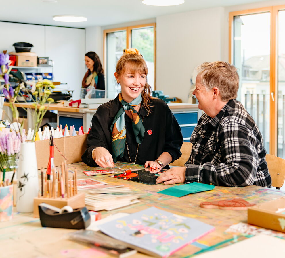 Eine Mitarbeiterin der Heilsarmee und ein Klient sitzen gemeinsam an einem Tisch und arbeiten an einem kreativen Projekt im Atelier an der Ankerstrasse in Zürich.