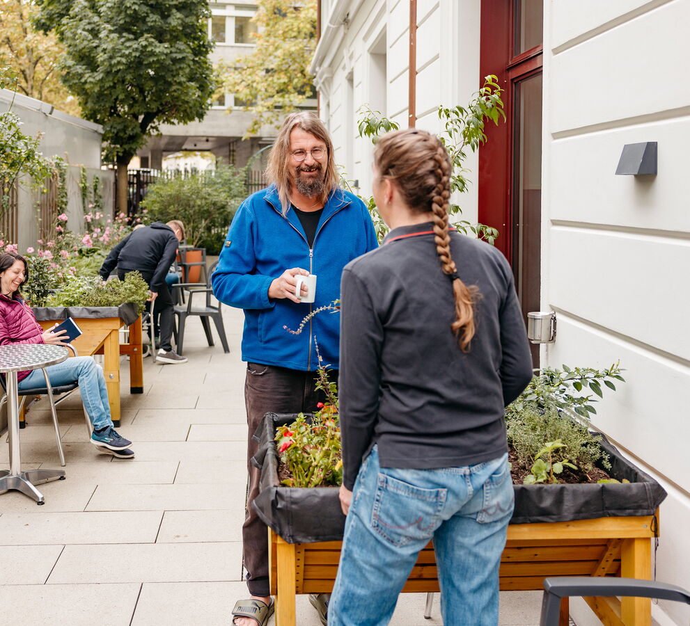 Ein Bewohner und eine Mitarbeiterin unterhalten sich bei einer Kaffeepause im begrünten Aussenbereich des Wohnhauses an der Molkenstrasse in Zürich. Im Hintergrund halten sich weitere Personen auf.