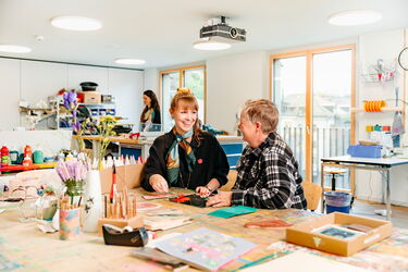 Eine Mitarbeiterin der Heilsarmee und ein Klient sitzen gemeinsam an einem Tisch und arbeiten an einem kreativen Projekt im Atelier an der Ankerstrasse in Zürich.